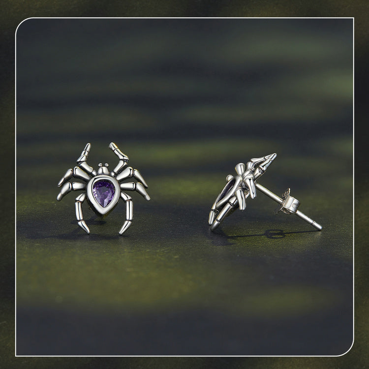 Silver spider-shaped earrings with a purple gemstone on a dark green background